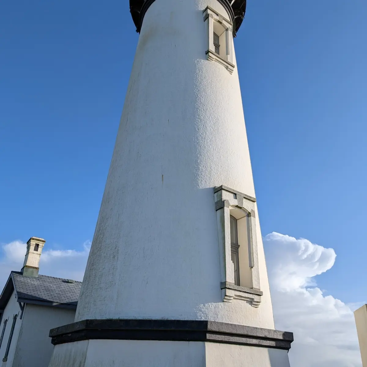A low-angle photo shows the white, textured tower of a lighthouse against a bright blue sky with scattered clouds. A small…
