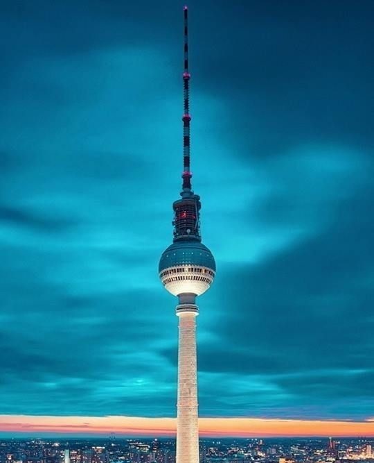 A tall, illuminated tower with a spherical observation deck and antenna stands against a dramatic blue and orange twilight…