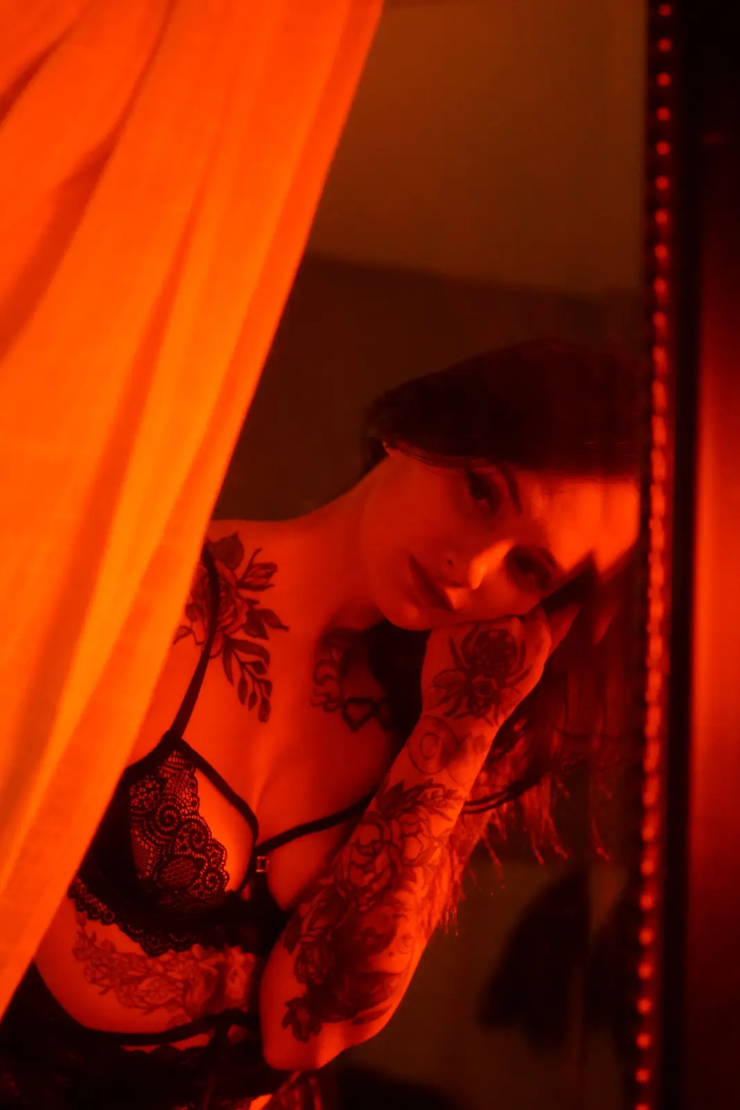 A woman with tattoos on her neck, chest, and arm, wearing black lace lingerie, looks out from behind a red curtain in a…