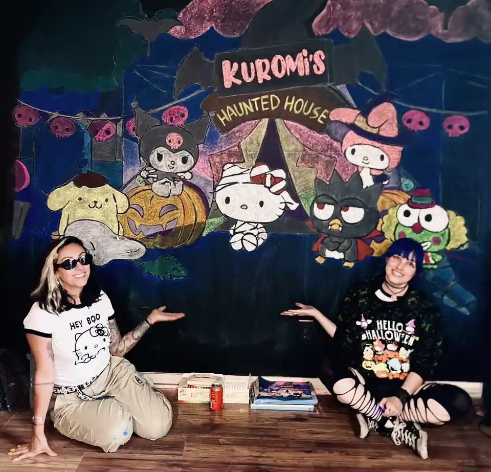 Two women sit on a wooden floor in front of a colorful chalkboard mural depicting a Halloween scene with Sanrio characters…