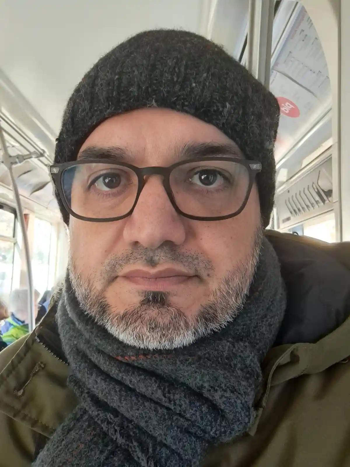 A man with glasses, a dark knit beanie, and a grey scarf looks directly at the camera while seated inside a public transit…