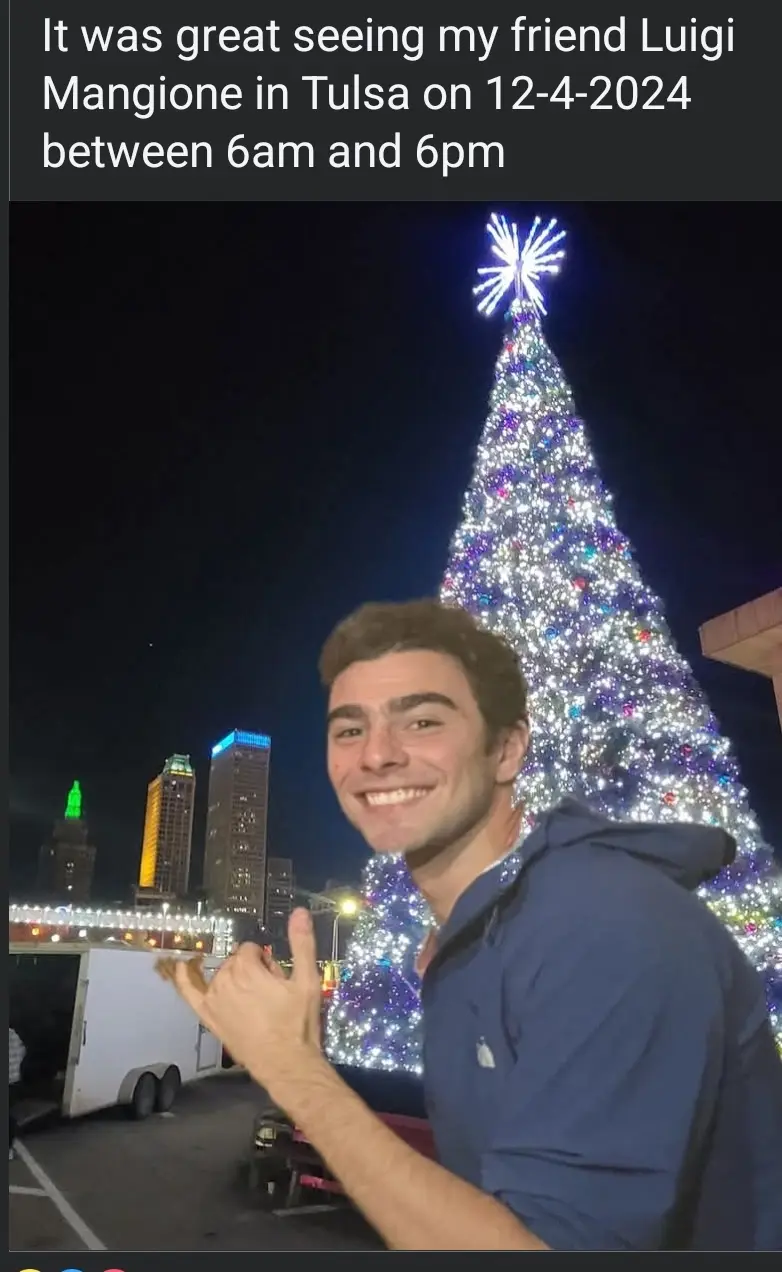 A smiling young man in a blue hoodie poses in front of a large, brightly lit Christmas tree at night, with city buildings…