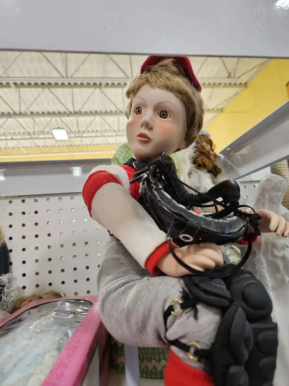 A doll with brown hair and a red hat is sitting on a store shelf, holding a pair of black roller skates. The doll wears a…