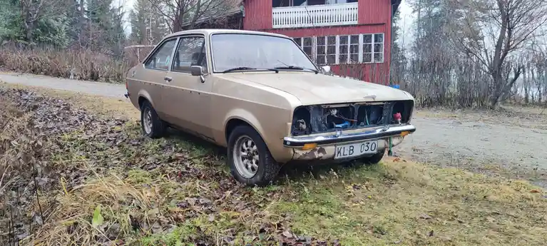 A beige, visibly aged and neglected vintage car with its front bumper and grille removed, exposing internal components, is…