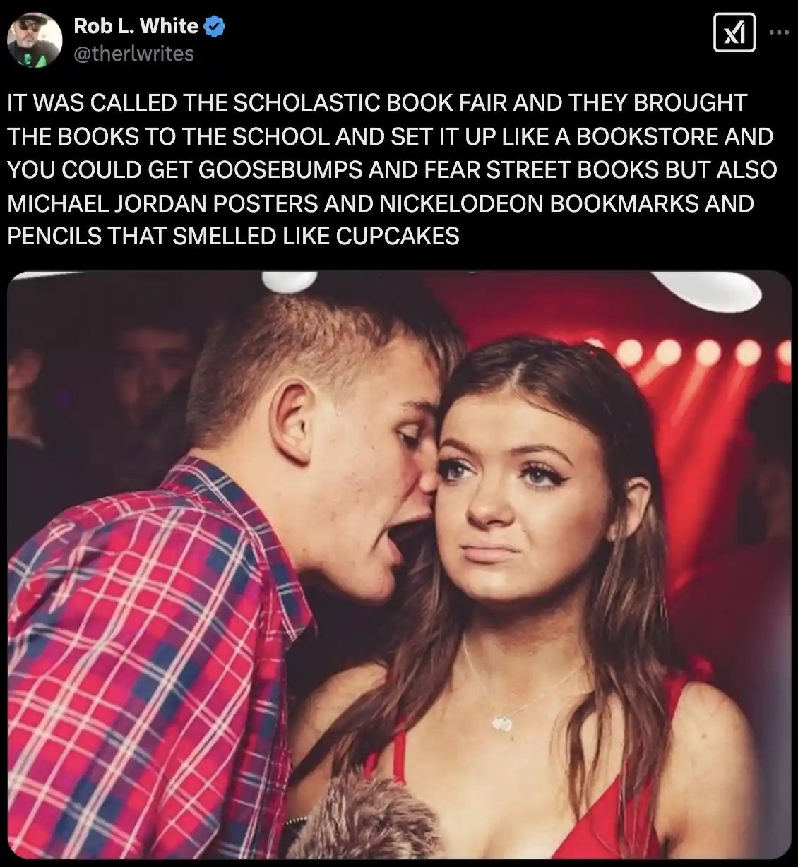 A young man in a red and blue plaid shirt whispers into the ear of a young woman in a red top, who looks unimpressed. The…