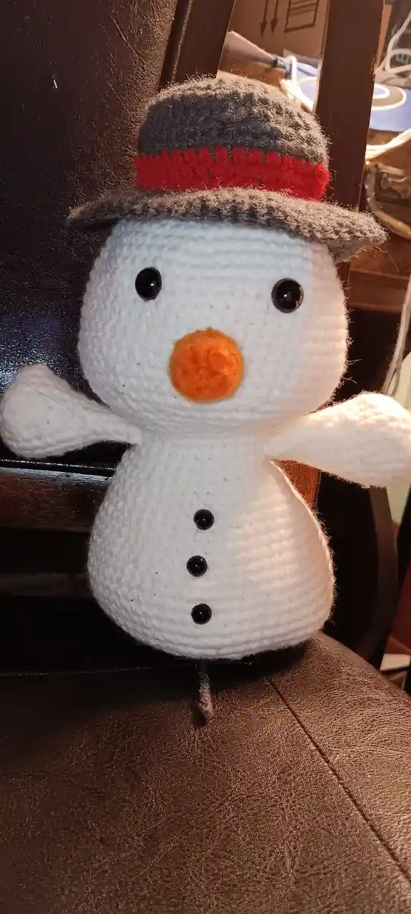 A handmade crocheted snowman plushie with black button eyes, an orange pom-pom nose, and three black buttons down its torso…
