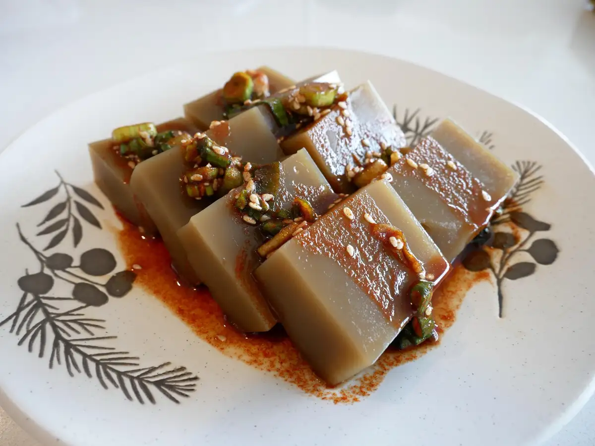 A plate of translucent, gelatinous cubes, likely Korean 'sannakji' or similar dish, topped with chopped green onions, sesame…