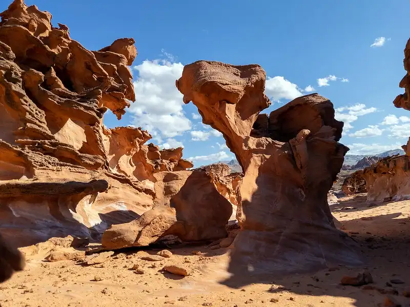 A desert landscape with orange-red sandstone formations under a blue sky with scattered clouds. The central rock formation…