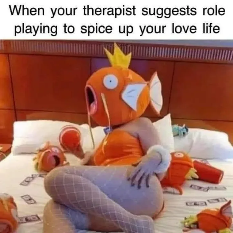 A person wearing an orange fish costume with a crown sits on a bed surrounded by other fish plush toys, looking shocked with…