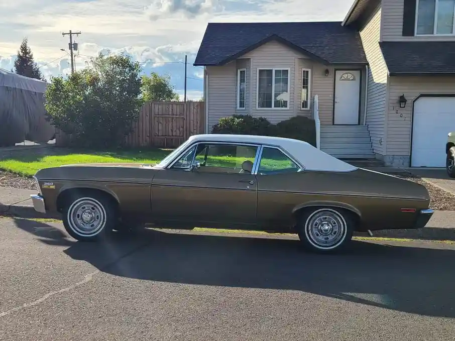 A brown and white classic American muscle car, likely a Chevrolet Chevelle, is parked on a residential street in front of a…