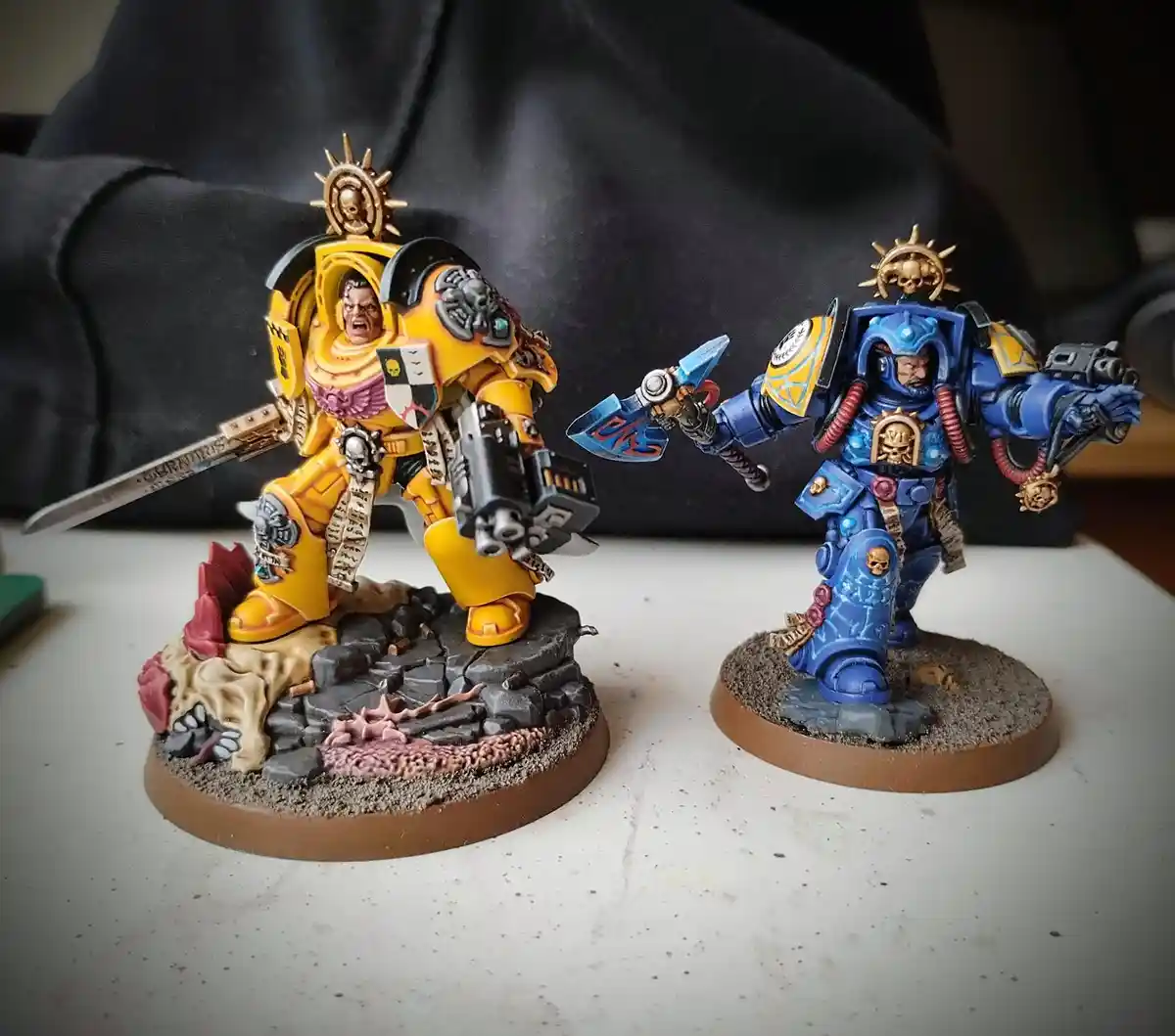 Two painted Warhammer 40,000 Space Marine miniatures stand side-by-side on a table; the one on the left is yellow and…