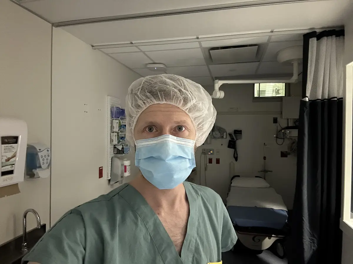 A man in green scrubs, a white surgical cap, and a blue surgical mask stares directly at the camera with wide eyes in a…