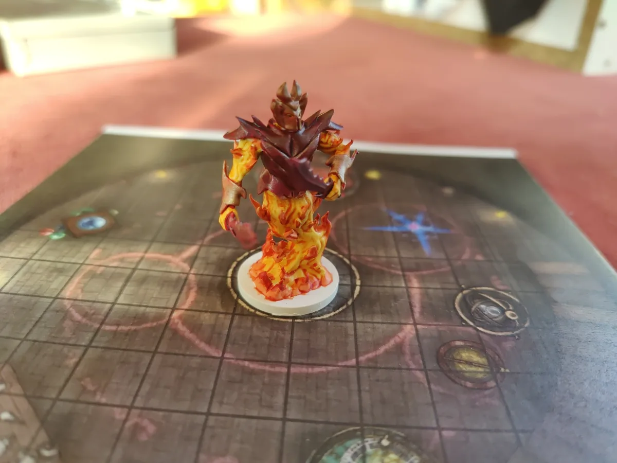 A painted miniature of a fire elemental, with orange and red flame-like textures and dark red spiky armor, stands on a white…