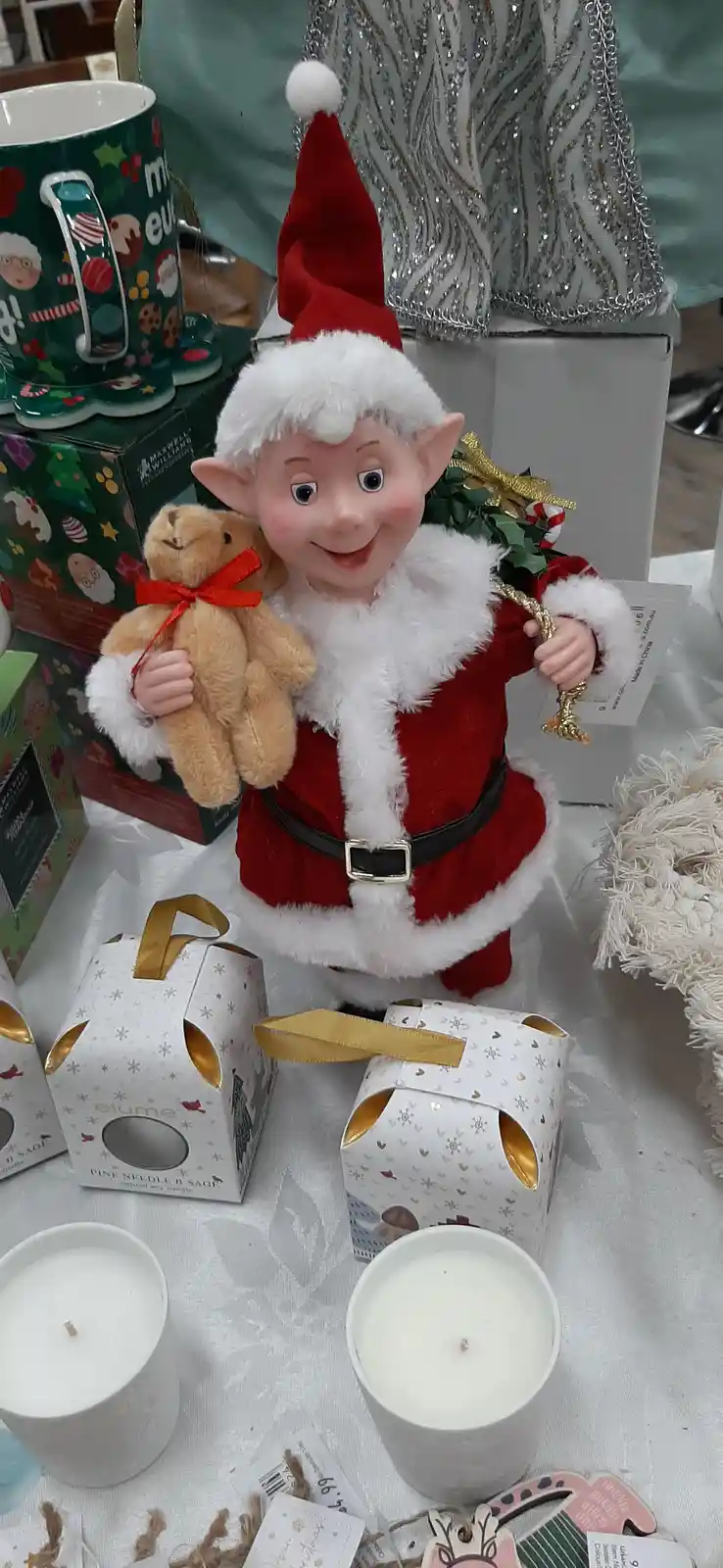 A plastic Christmas elf doll dressed in a red and white Santa outfit, holding a small teddy bear and a holly branch, is…