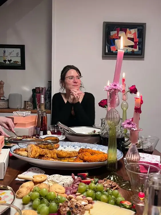 A woman sits at a lavishly set dinner table with abundant food, including fried items, cheese, grapes, and a large platter…