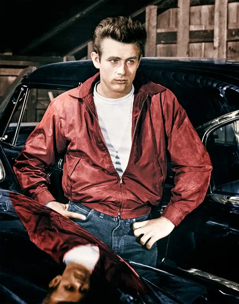 a man in a red jacket and jeans leans against a black car, looking intensely at the camera. His reflection in the car's…