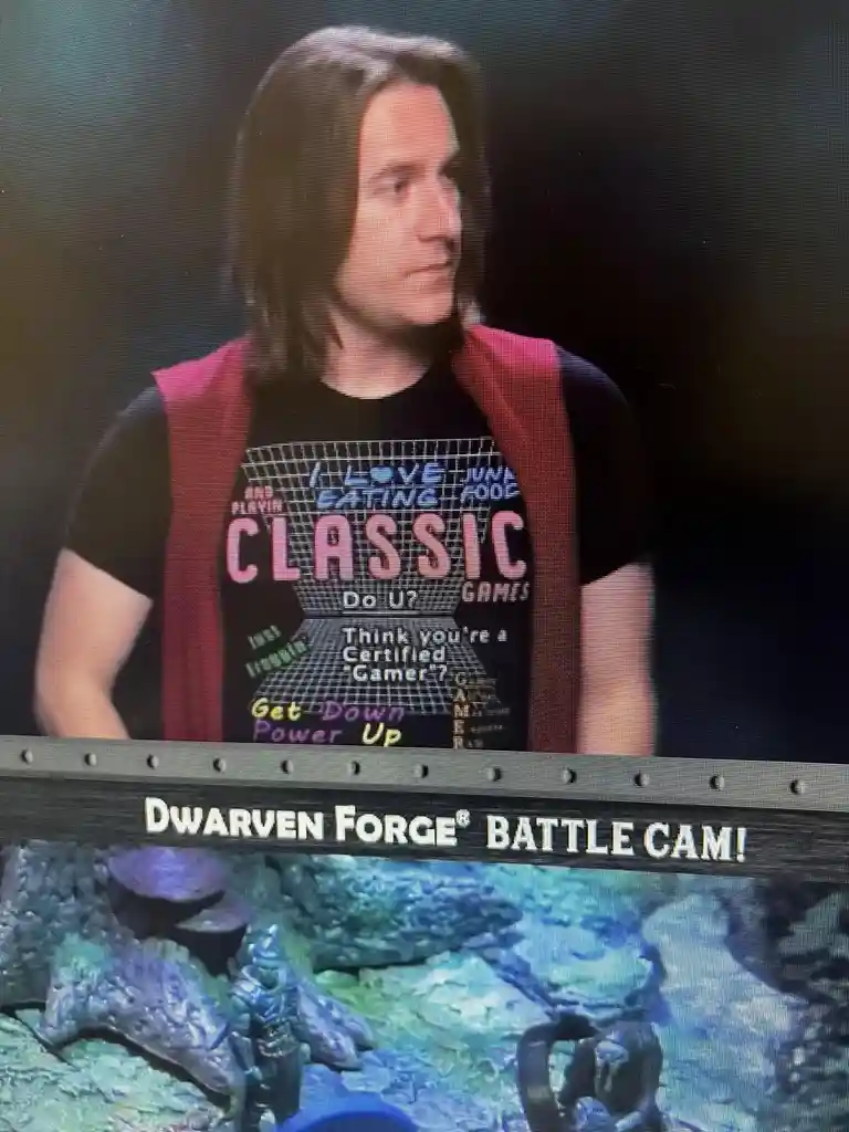 A man with long dark hair wearing a black t-shirt with retro gaming text and a red vest is shown on a screen. The t-shirt…