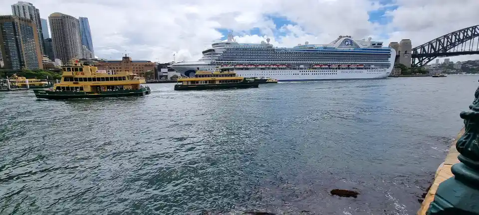 A wide shot of Sydney Harbour shows a large white Princess Cruises ship named 'Crown Princess' sailing past yellow and green…