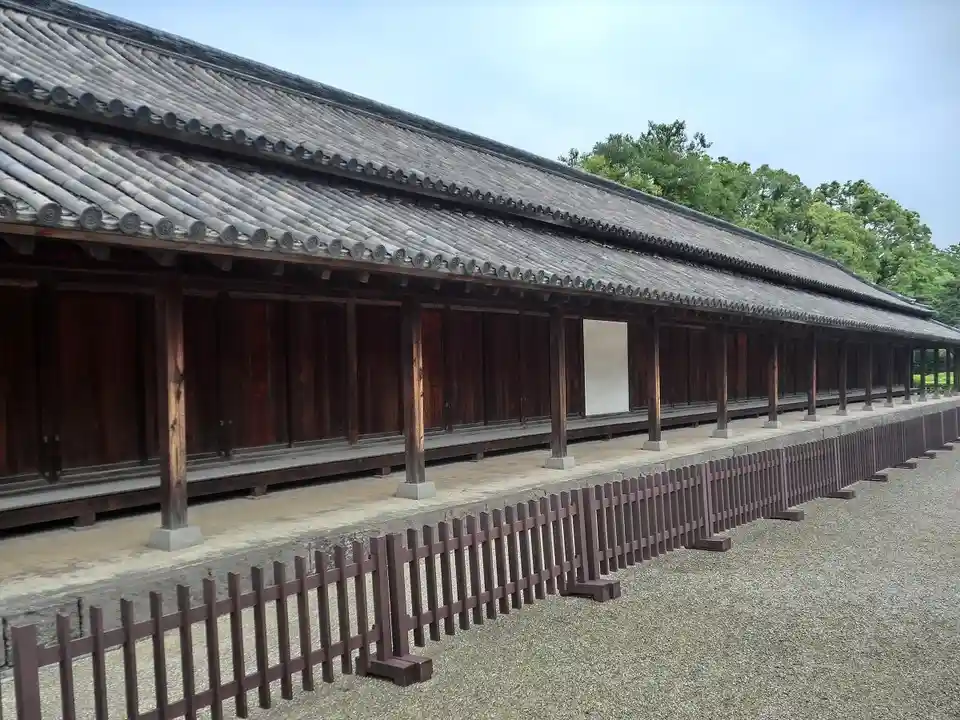 A long, traditional wooden building with a tiled roof and a low fence in front, under a clear sky. The overlaid text reads…