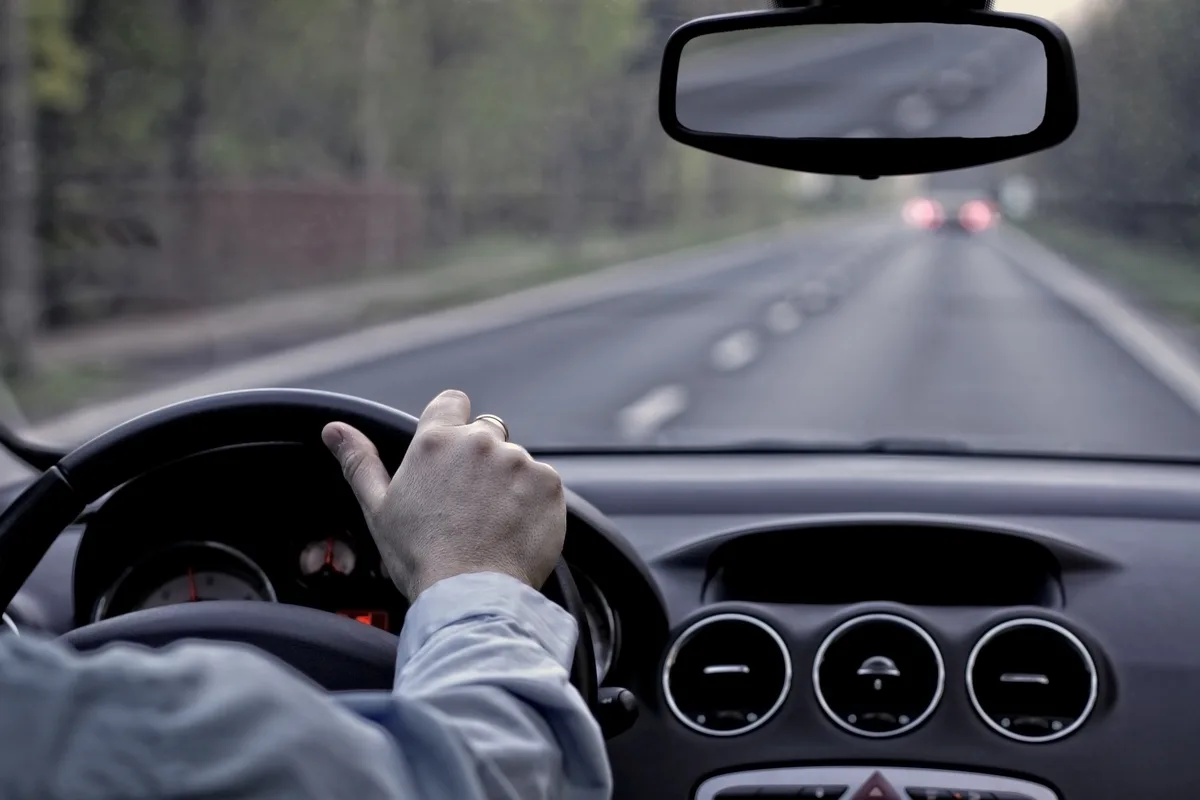 A first-person view from inside a car shows a driver's hand on the steering wheel, with a blurry road ahead and brake lights…