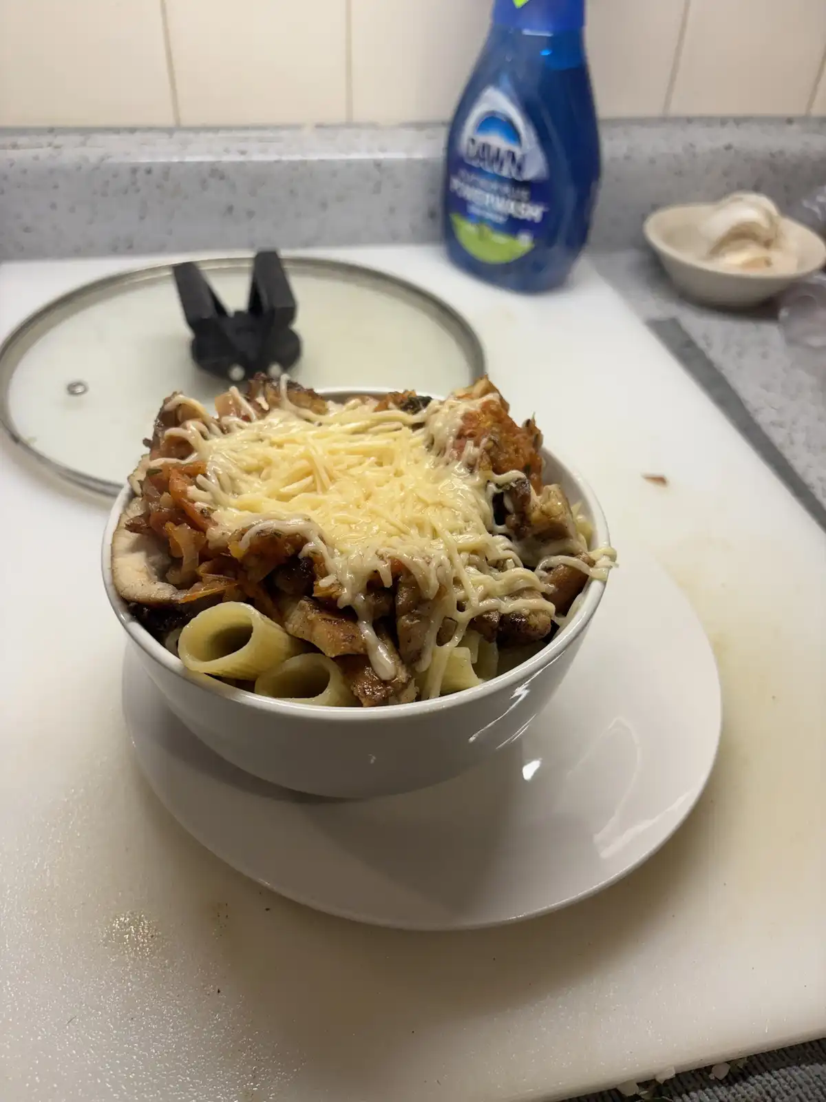 A white bowl filled with pasta, meat, and melted cheese sits on a white plate on a kitchen counter. In the background, a…