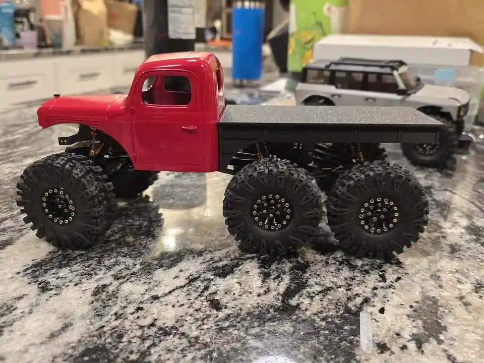 A red remote-controlled flatbed truck with oversized, knobby tires sits on a granite countertop, with a silver RC SUV…