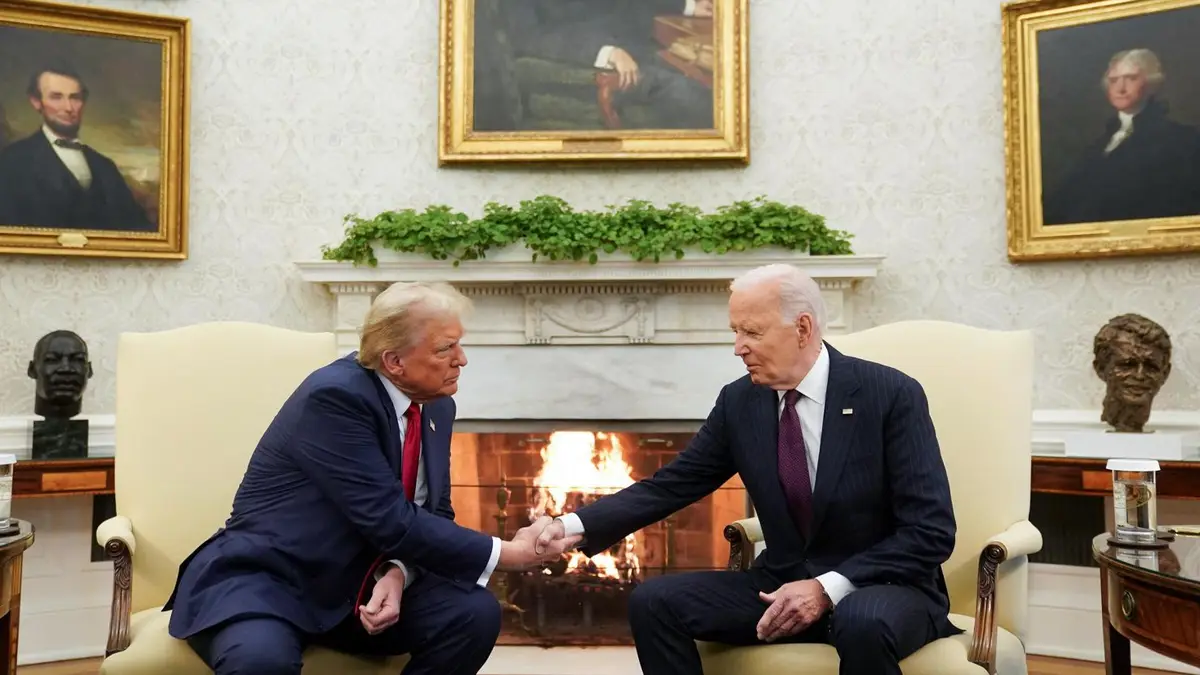 Two men in suits sit in chairs facing each other across a fireplace in an ornate room with framed portraits on the wall. One…