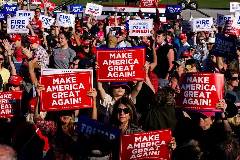 A crowd of people at a political rally holds up red signs with 'MAKE AMERICA GREAT AGAIN!' and some also hold blue signs…