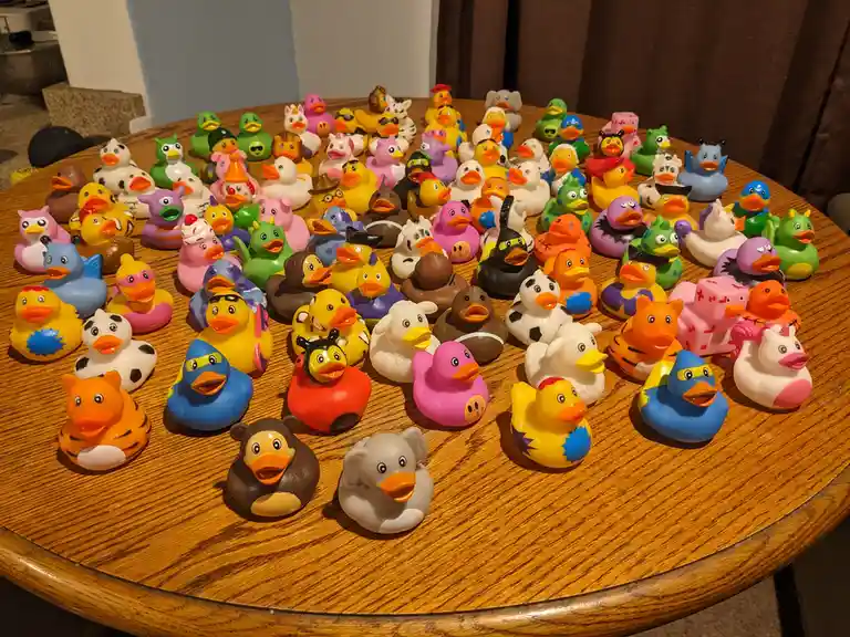 A large wooden table is covered with dozens of colorful rubber ducks, many customized with different designs, colors, and…