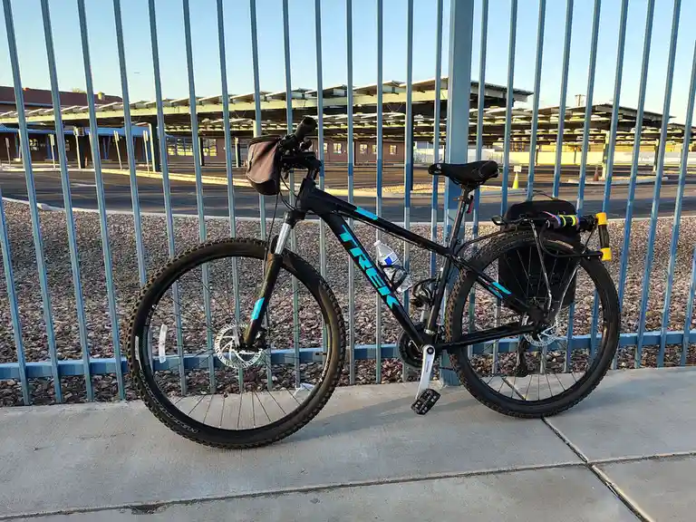 A black Trek mountain bike with blue accents is parked against a blue metal fence on a sidewalk, equipped with a water…