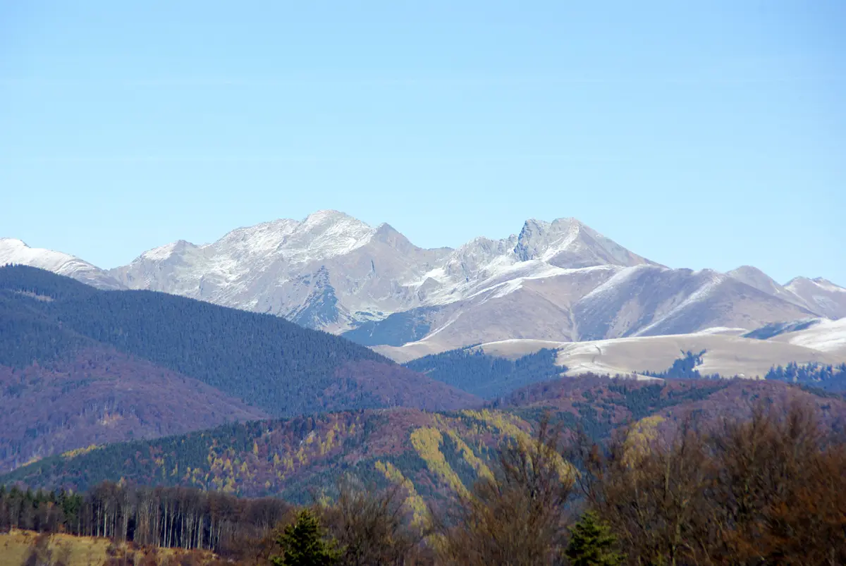 A scenic mountain landscape with snow-capped peaks under a clear blue sky, layered with forested hills showing autumn…