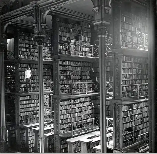 A black-and-white photo shows a person standing on a narrow walkway between towering, multi-level bookshelves in a grand…