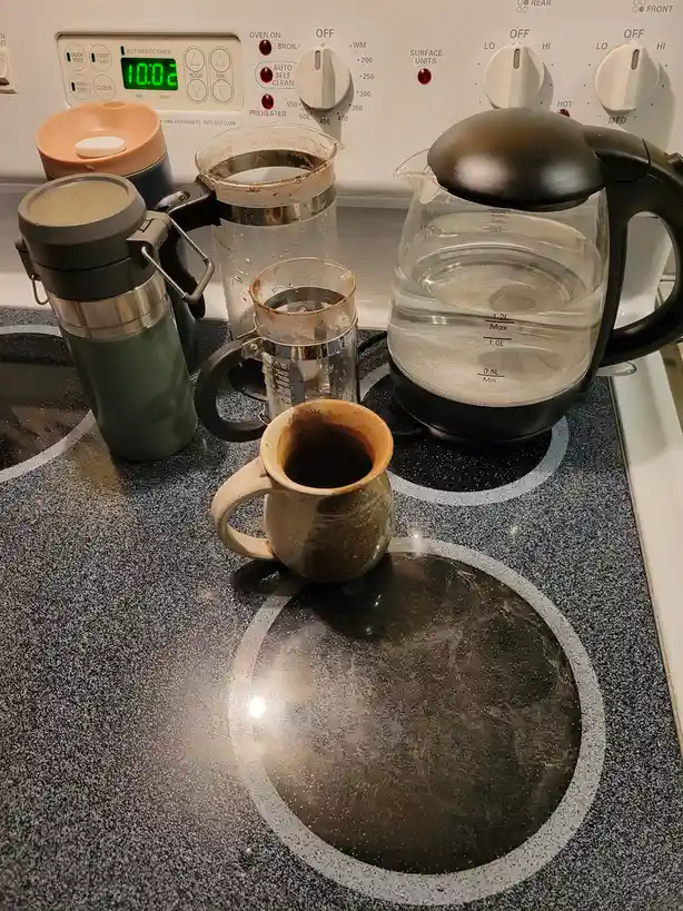 An electric stove top with a digital clock showing 10:02, surrounded by coffee-making equipment including a French press, a…