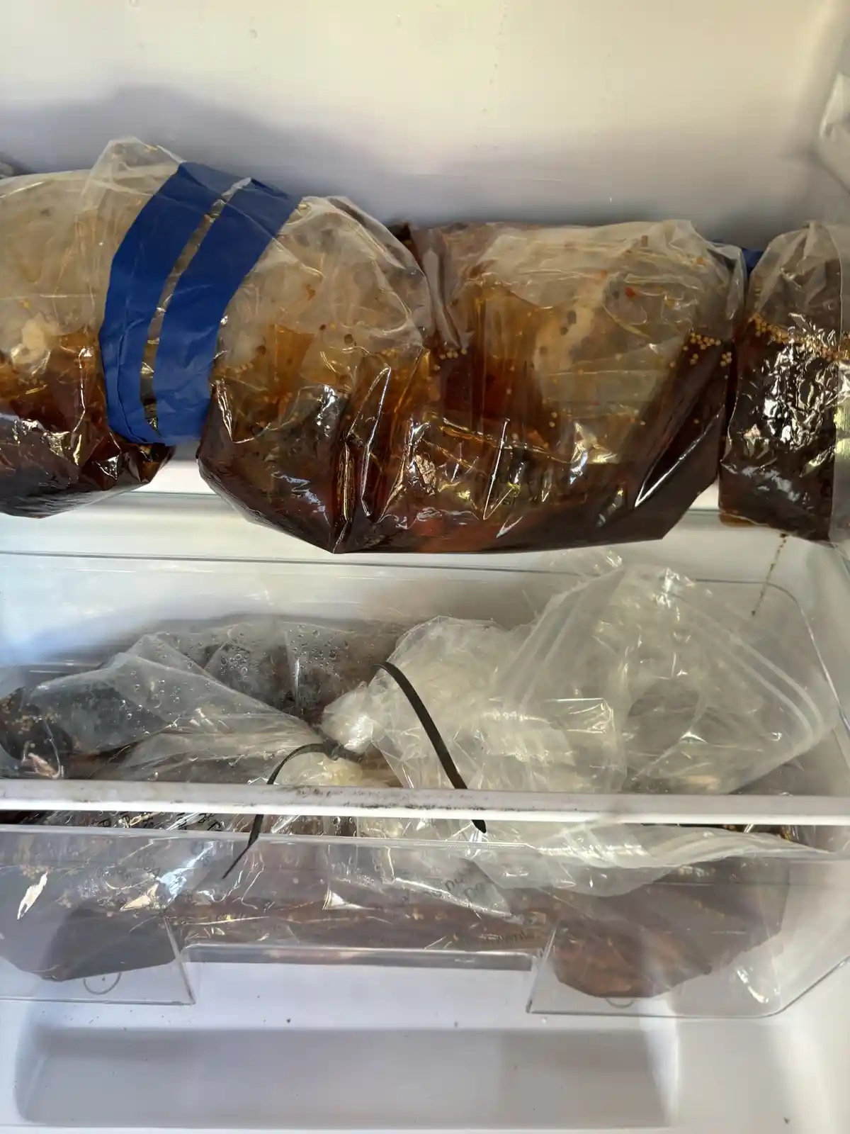 A freezer contains multiple clear plastic bags filled with dark brine and chicken pieces, some sealed with blue tape and…