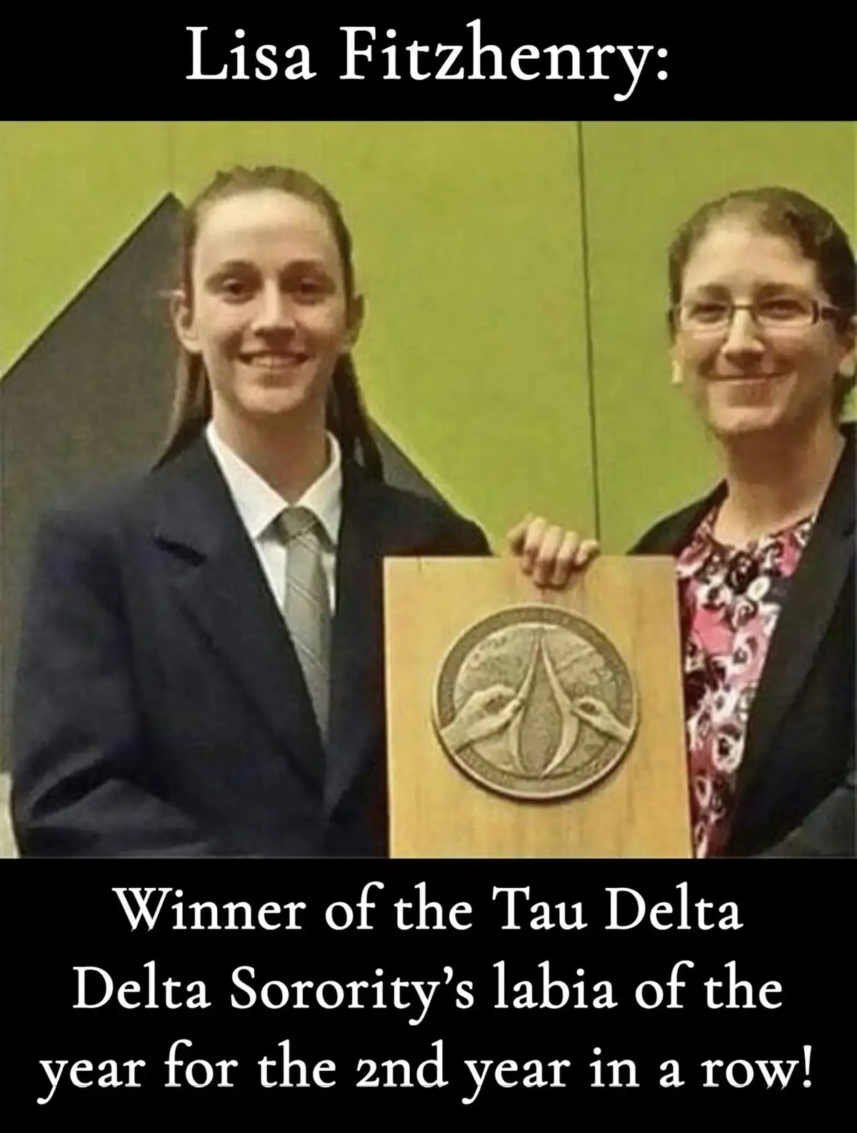 Two women stand smiling while holding a wooden plaque with a circular emblem. The text at the top identifies the younger…