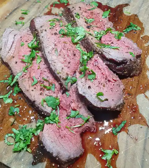 Sliced medium-rare steak with a pink center and seared edges, garnished with chopped cilantro and black pepper, rests on a…