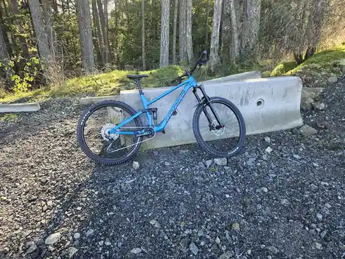 A bright blue Norco mountain bike is leaning against a concrete barrier on a gravel path in a forest. The bike has black…
