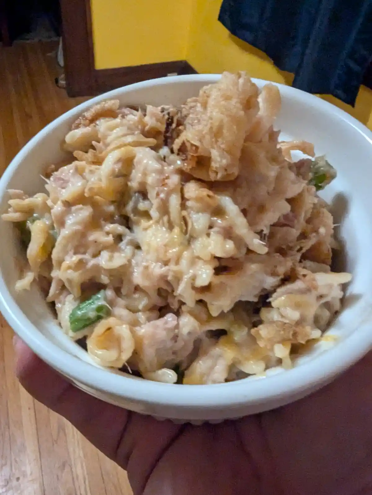A close-up photo shows a hand holding a white bowl filled with a creamy, cheesy pasta dish topped with crispy fried onions…