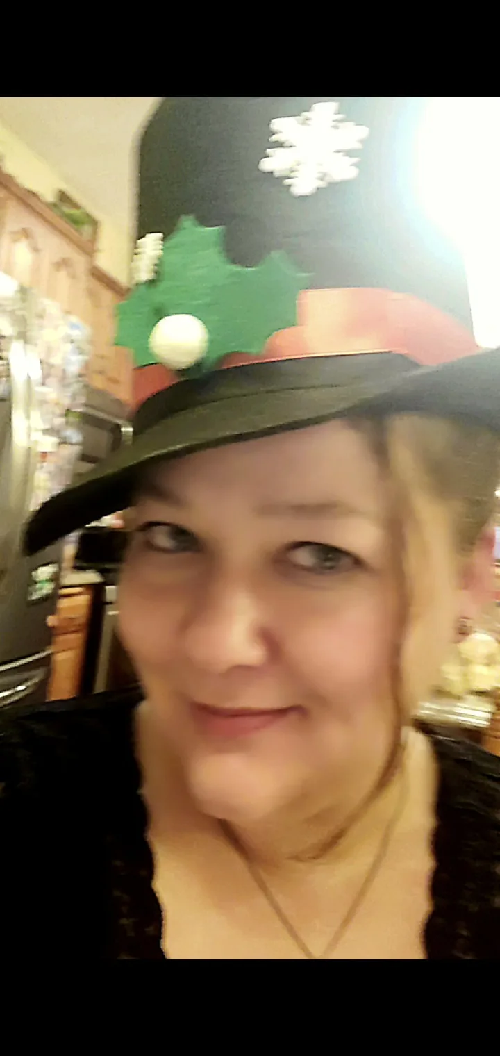 A blurry selfie of a woman with blonde hair wearing a tall, decorated Christmas hat featuring a green holly leaf with a…