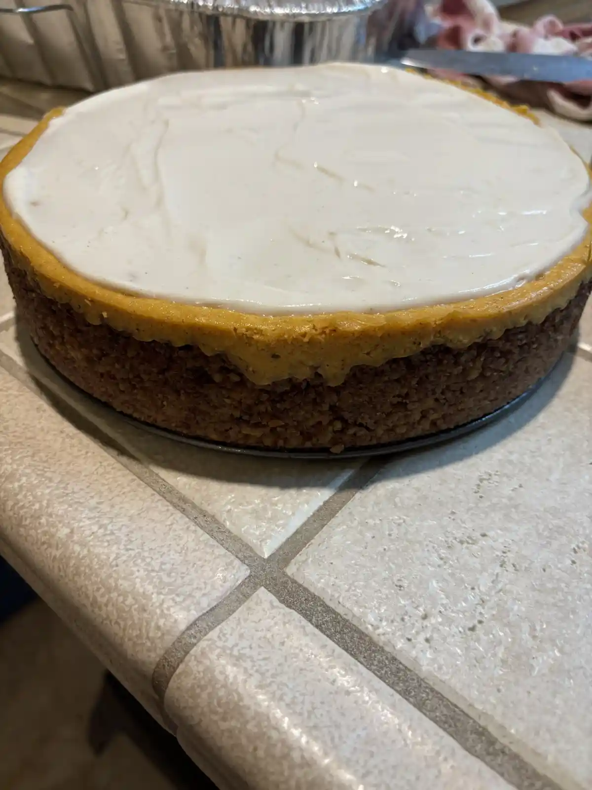 A close-up photo shows a round pumpkin cheesecake with a golden-brown crust and creamy white topping, sitting on a metal pan…