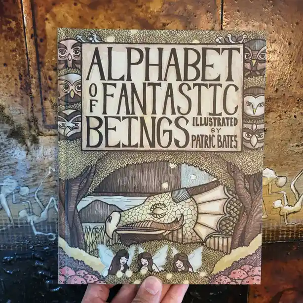 A hand holds a children's book titled 'Alphabet of Fantastic Beings' illustrated by Patric Bates. The cover features a…