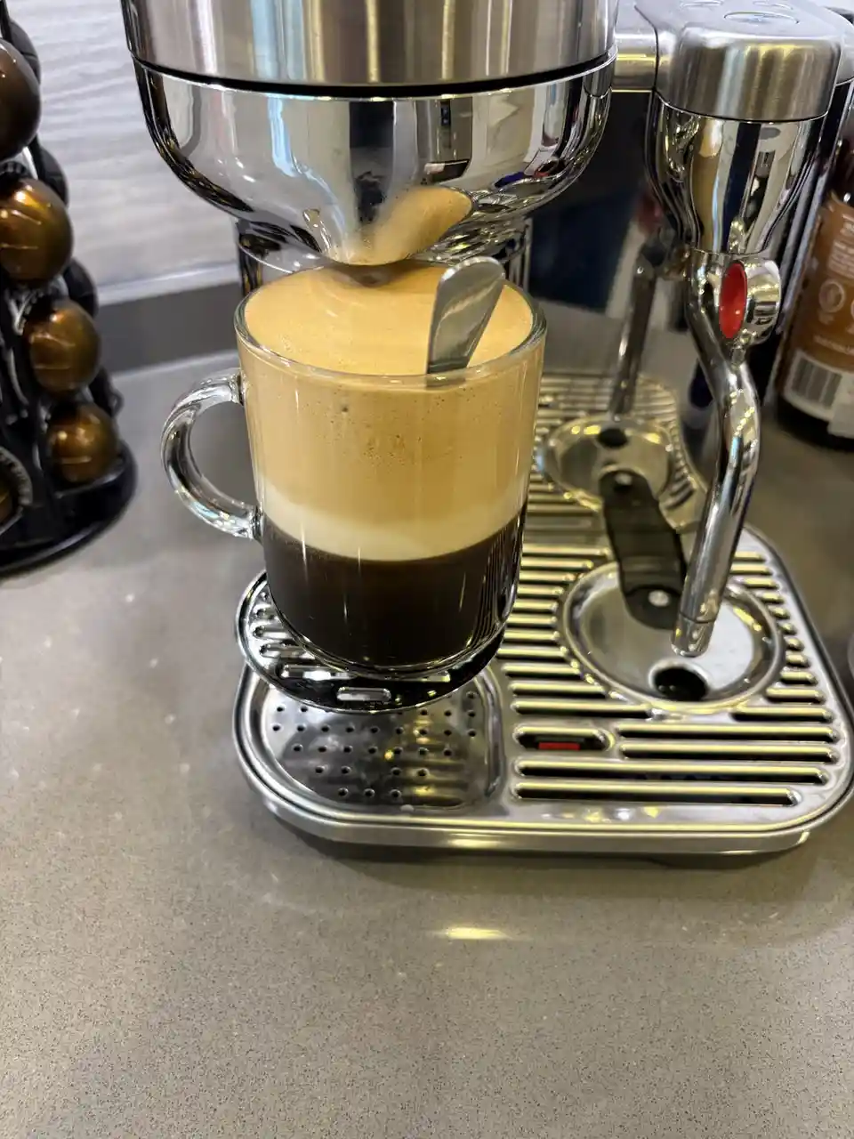 A clear glass mug filled with layered coffee sits under a stainless steel espresso machine, with a spoon resting in the…