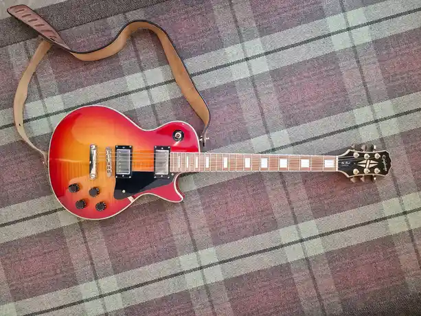 An electric guitar with a sunburst finish and a brown leather strap lies on a plaid blanket. The guitar has a glossy…