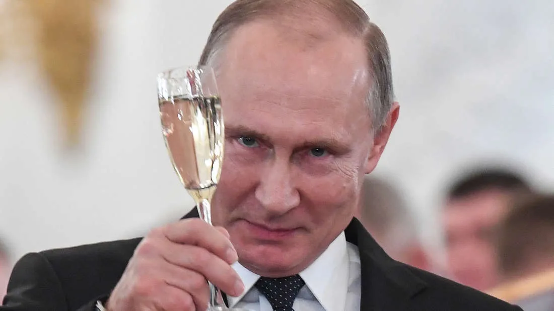 a close-up photo of Vladimir Putin holding a champagne flute, looking intently at the camera with a serious expression. He…