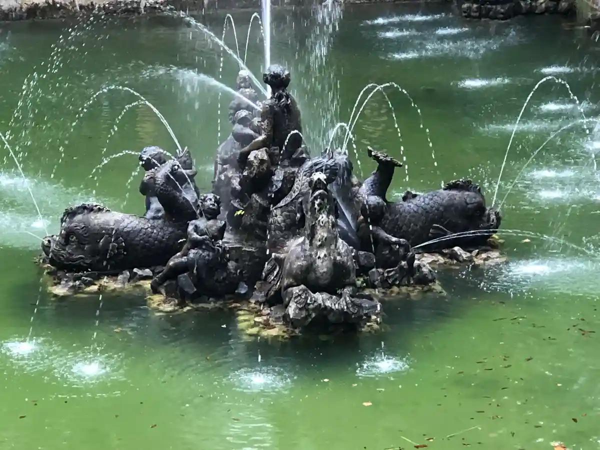 A dark bronze fountain sculpture with multiple figures and fish-like creatures, all spouting water into a greenish pond. The…