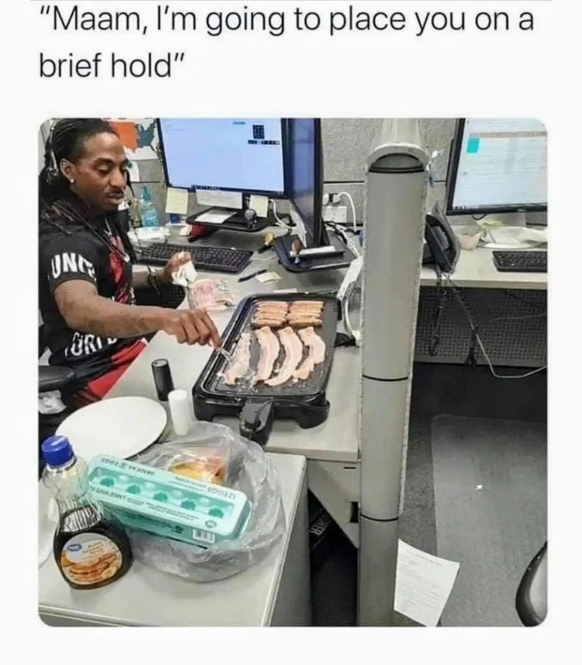 A man wearing headphones sits at an office desk with multiple monitors, using a small electric grill to cook bacon and…