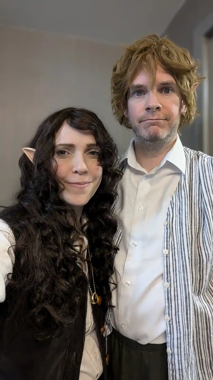 A man and woman stand side-by-side, both wearing pointed elf ears. The woman, on the left, has long dark curly hair and a…