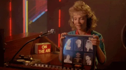 A woman with curly blonde hair and headphones sits at a radio station desk with a microphone and an 'ON THE AIR' sign. She…