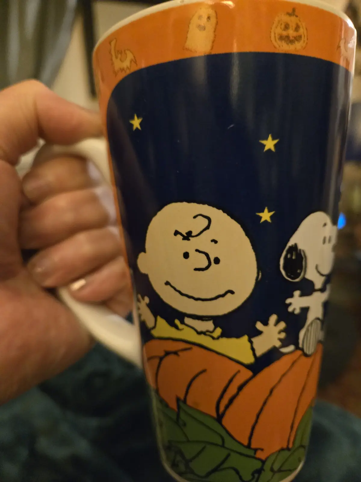 A person holds a Halloween-themed mug featuring Charlie Brown and Snoopy inside a pumpkin under a starry night sky. The…