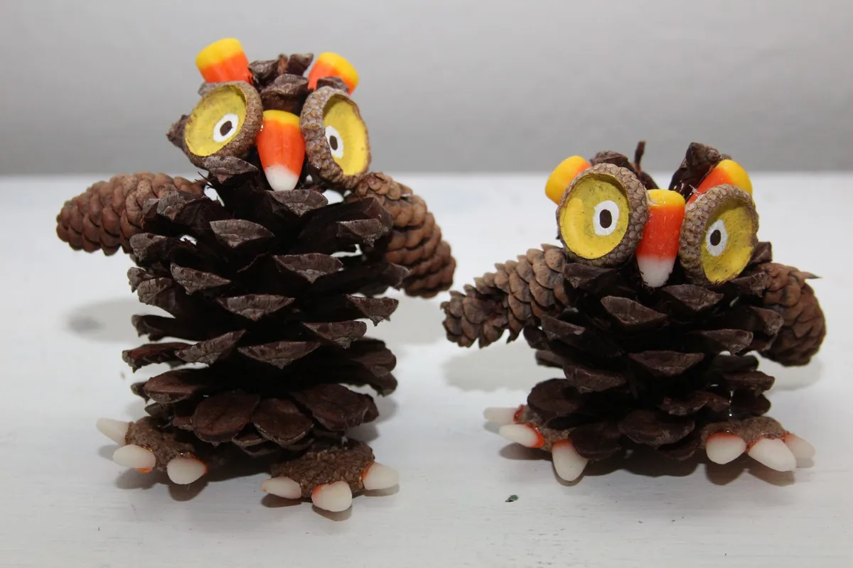 Two handmade owl crafts made from pinecones, with acorn caps as eyes, candy corn for beaks and ears, and white plastic…